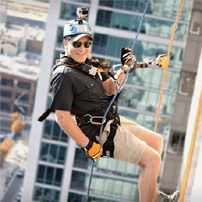 The image shows a man rappelling down the side of a tall building. He is wearing a helmet, sunglasses, and safety gear. He appears to be smiling and enjoying the experience. The background shows other buildings, suggesting an urban environment.